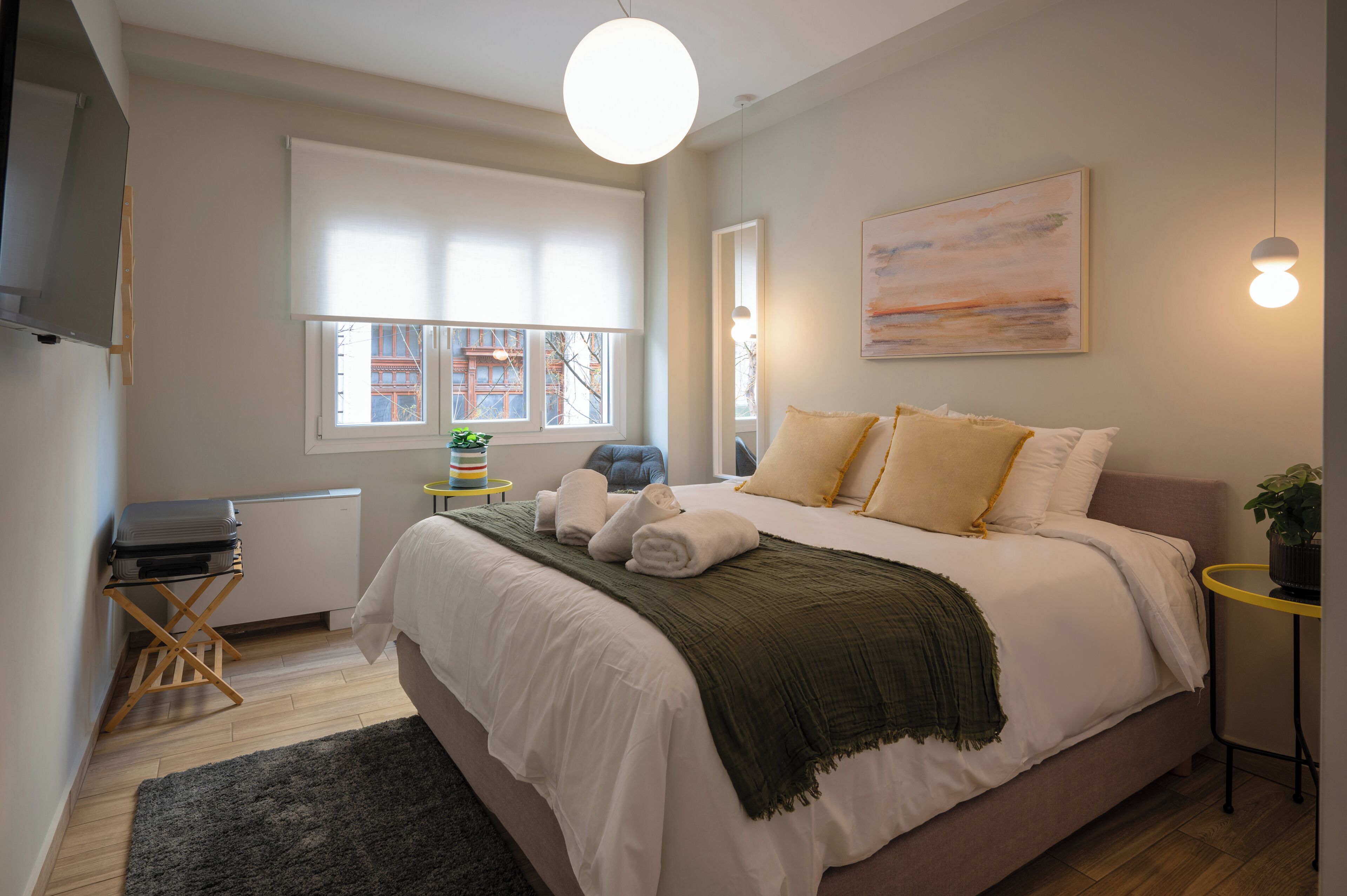 A bedroom with a bed, a window, a radiator, a small table, and a plant.