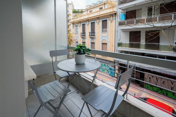 A balcony with a round table and two chairs, a potted plant, and a view of a building with balconies and graffiti.