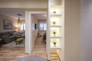 A modern living room with a sofa, coffee table, and pendant lights. There is a hallway leading to another room with a bed and a window.