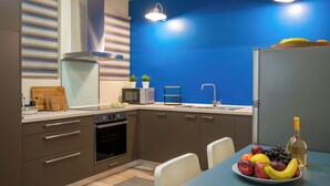 A modern kitchen with a blue accent wall, stainless steel appliances, a dining area with a fruit bowl, and a bottle of wine. - Mia Homes - National Museum, Mia Homes (Athens)
