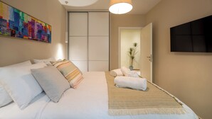 A hotel room with a bed, pillows, a blanket, a hanging lamp, a painting, a potted plant, and a TV on the wall. - Mia Homes - National Museum, Mia Homes (Athens)
