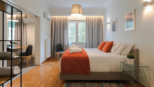 Une chambre à coucher moderne avec un lit, une table de chevet en verre, une chaise, un bureau et une fenêtre avec des rideaux.