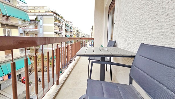A balcony with a table and chairs, overlooking a street with buildings and parked cars.