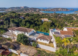 A residential area with houses featuring tiled roofs and solar panels, surrounded by lush greenery and a coastal view. - Bz72 Casa Vista Mar 5 suítes e piscina (Buzios)