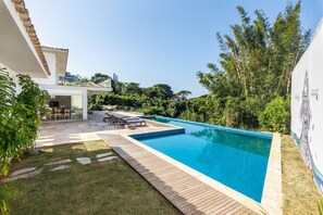 A swimming pool with a wooden deck, surrounded by greenery and a white building with a tiled roof. - Bz72 Casa Vista Mar 5 suítes e piscina (Buzios)