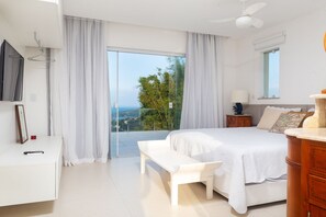 A bedroom with a bed, a bench, a bedside table, a TV, and a view of the outdoors. - Bz72 Casa Vista Mar 5 suítes e piscina (Buzios)
