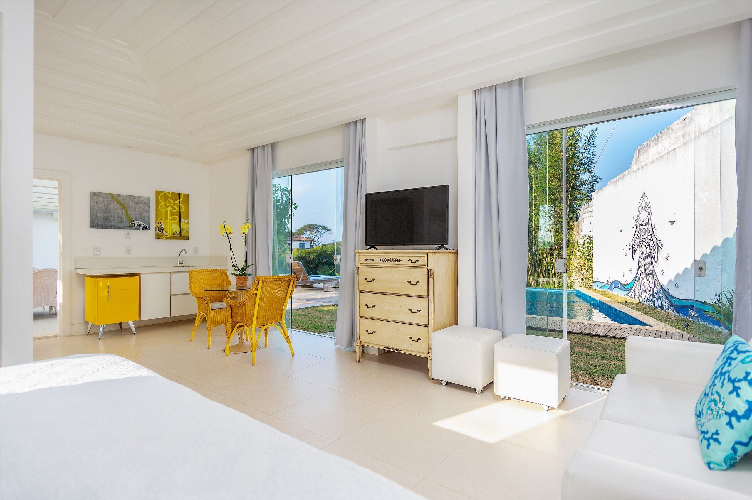 A modern hotel room with a large bed, a dining area with yellow chairs, a white sofa, and a TV.