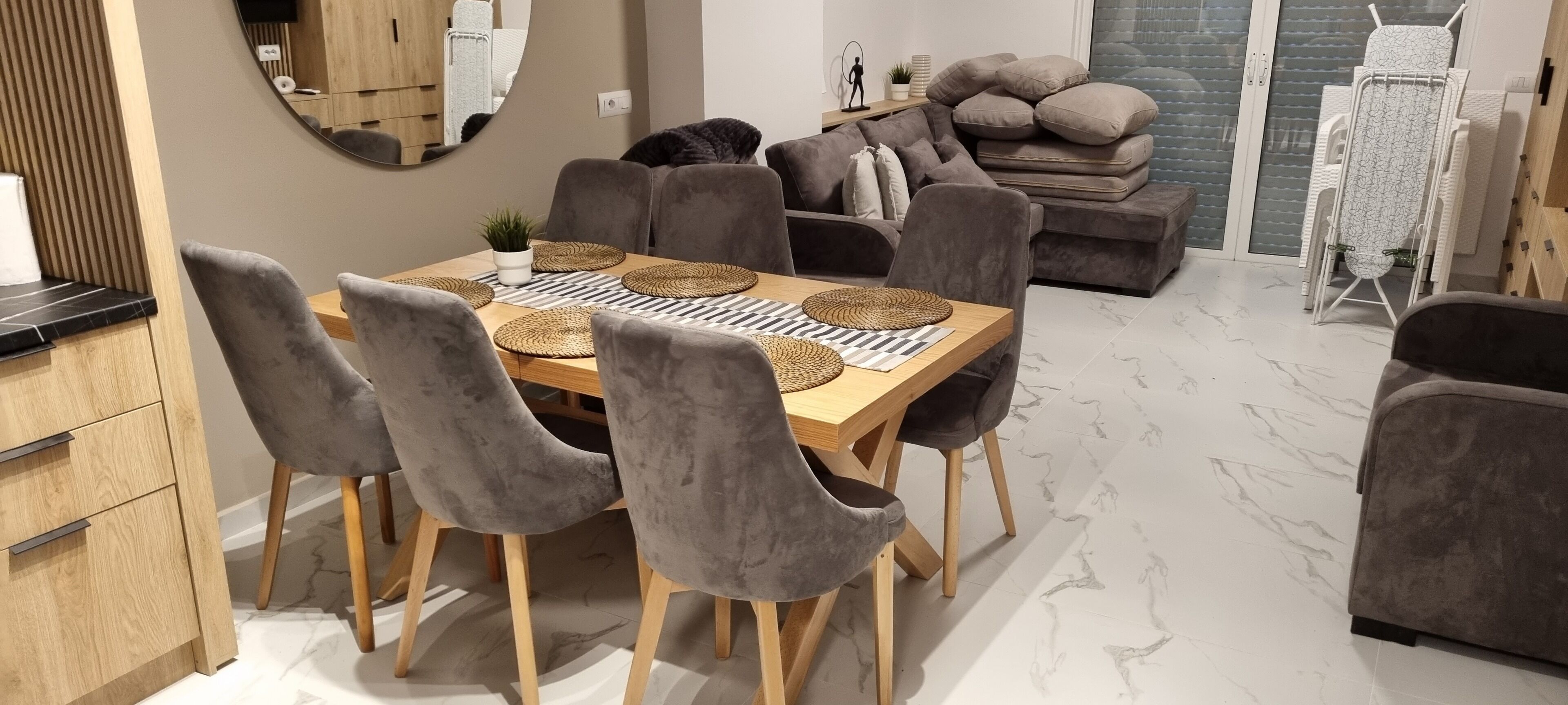 Ein stilvoller Essbereich mit einem Holztisch, grau gepolsterten Stühlen, einer grauen Couch und einem Kissenstapel.