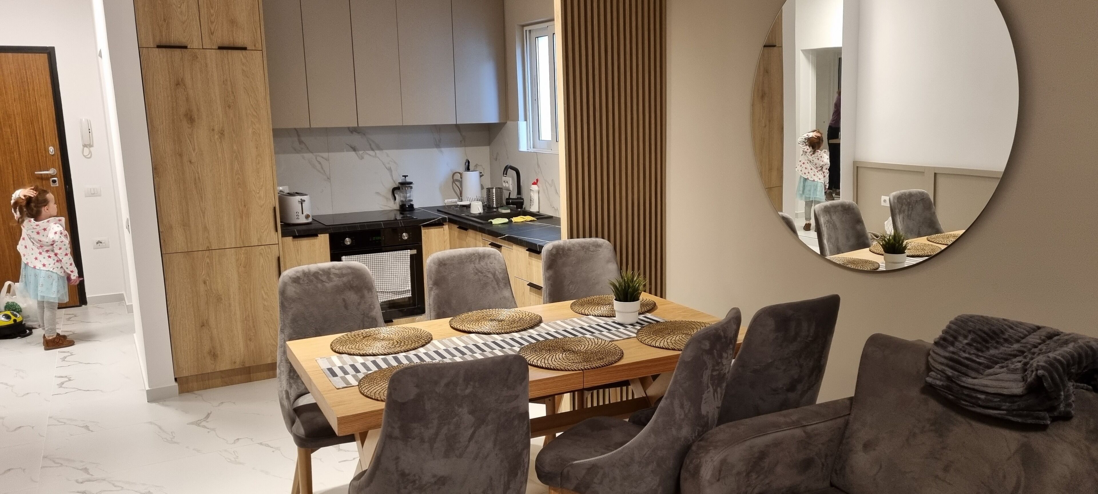 Eine moderne Küche mit Holdekor und einer schwarzen Arbeitsplatte, ein Essbereich mit einem Holztisch und grau gepolsterten Stühlen sowie ein großer Spiegel an der Wand.