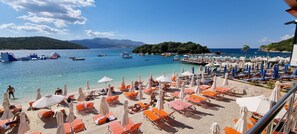 A beach with numerous sun loungers and umbrellas, boats on the water, and a mountainous backdrop.