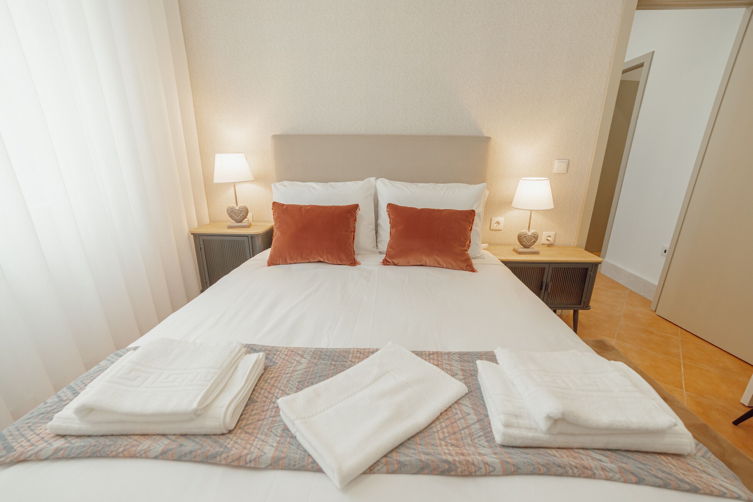 A neatly made bed with white linens, two red pillows, and two bedside lamps with heart-shaped lampshades.