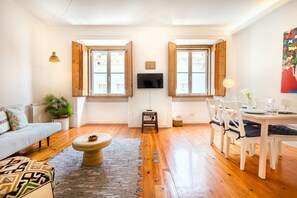 A living room with a sofa, coffee table, dining table with chairs, a TV, and a painting on the wall. - Central Apartment at Chiado (Lisbon)