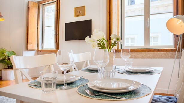 A dining table set with plates, glasses, and a vase of flowers, with a TV mounted on the wall and a window with a view of buildings. - Central Apartment at Chiado (Lisbon)