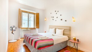 A bedroom with a large bed, a bedside table with a lamp, a window with wooden shutters, and a wall-mounted light fixture. - Central Apartment at Chiado (Lisbon)