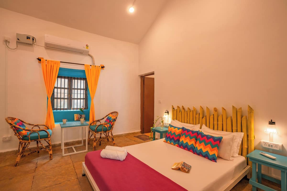 A room with a bed, a table, two chairs, a window with curtains, an air conditioning unit, and a bedside lamp.