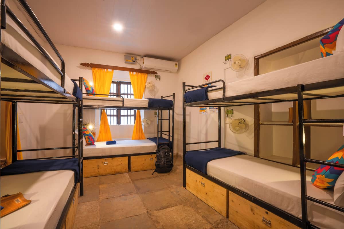 A dormitory room with bunk beds, a window with curtains, an air conditioning unit, and a fan on the wall.