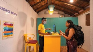 Une salle d’attente avec un plafond en bois, un bureau jaune et une chaise. On peut voir des panneaux sur le mur, ainsi qu’une personne portant un t-shirt jaune et une visière de protection.