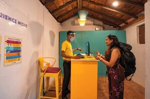 A reception area with a wooden ceiling, a yellow desk, and a chair. There are signs on the wall and a person wearing a yellow shirt and a face shield. - goSTOPS Lite Goa, Calangute (Calangute)