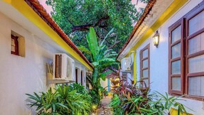 A narrow, tree-lined corridor with a stone pathway, flanked by buildings with yellow and white walls and wooden doors. - goSTOPS Lite Goa, Calangute (Calangute)
