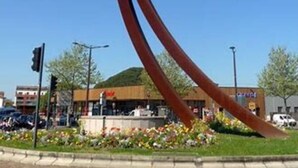 Een groot metalen beeld in een openbare ruimte, met een gebouw op de achtergrond.