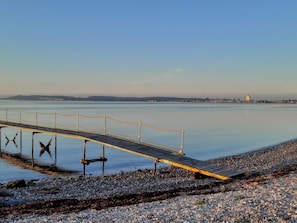 Una orilla tranquila con un muelle de madera que se adentra en el agua, rodeada de piedras y un horizonte urbano distante.