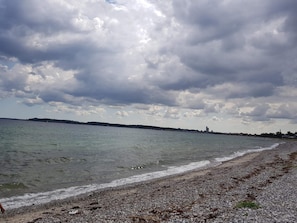 Una playa con orilla pedregosa, agua tranquila y una costa distante con edificios.