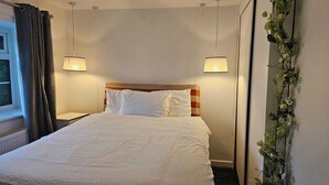 Ein Hotelzimmer mit einem Bett, zwei Nachttischlampen, einem fensterförmigen Ausblick mit Vorhängen und einer Wandpflanze.
