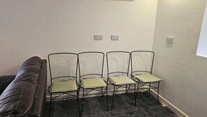 Ein Zimmer mit einem schwarz-weißen Fleckenmuster-Teppich, einem braunen Sofa und vier Metallstühlen mit grünen Kissen. An der Wand befinden sich zwei Steckdosen.