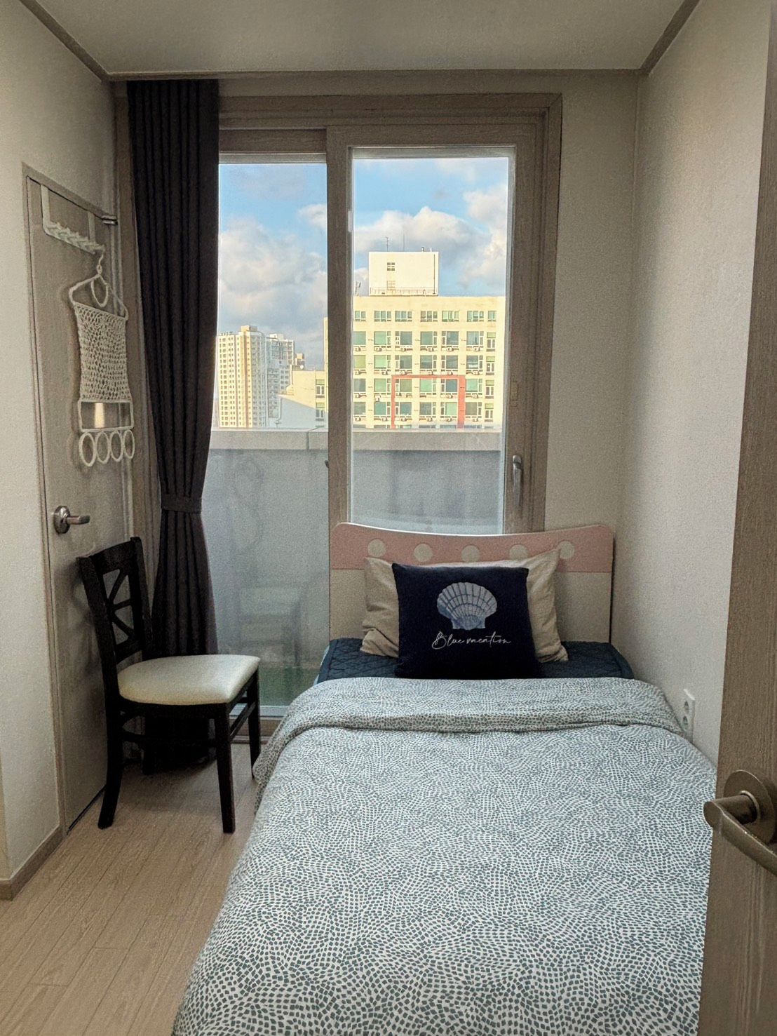 Un dormitorio con cama, una silla, un ventanal con vistas a edificios y una puerta.