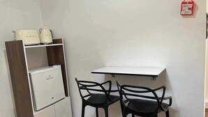 A small kitchenette with a refrigerator, a microwave, and a coffee maker. There are two black chairs and a white table. A red fire extinguisher is mounted on the wall. - Studio in a house (Sétif)
