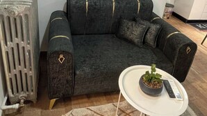 A living room with a dark grey sectional sofa, a small white side table with a plant, a radiator, a painting of a pink tree, and a clock on the wall. - Studio in a house (Sétif)