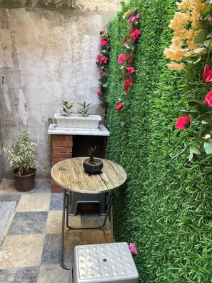 A small outdoor seating area with a round wooden table and metal chairs, a potted plant, and a green wall with flowers. - Studio in a house (Sétif)