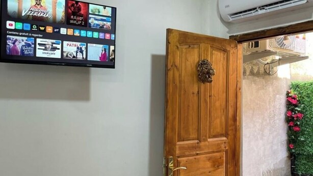 A room with a wooden door, a mounted television displaying various channels, and an air conditioning unit on the wall. - Studio in a house (Sétif)