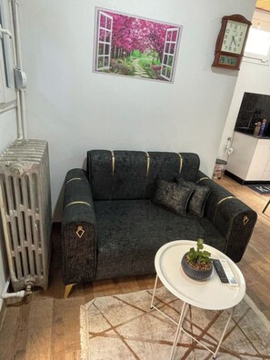 Un salon avec un canapé d’angle gris foncé, une petite table d’appoint blanche ornée d’une plante, un radiateur, un tableau représentant un arbre rose et une horloge murale.