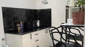A kitchen with white cabinets, black countertops, and a black backsplash. There is a 'Kitchen' sign on the wall, a potted plant, and a doormat with 'Kitchen' written on it. - Studio in a house (Sétif)