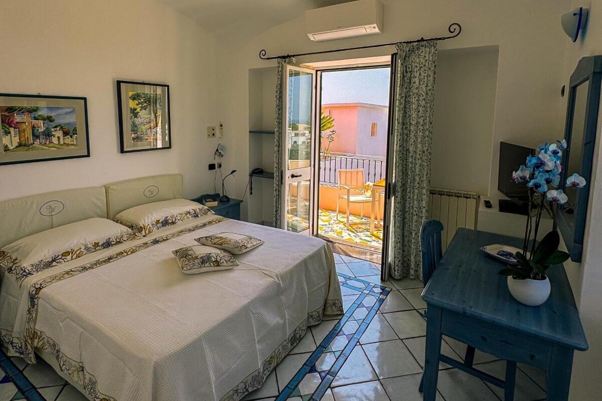 Un dormitorio con cama, ventilador de techo, escritorio con silla y vistas a un área exterior por una puerta de vidrio.