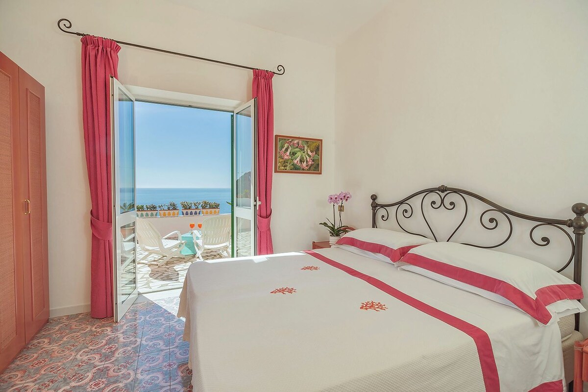A bedroom with a bed, a balcony with a view of the sea, and a door.