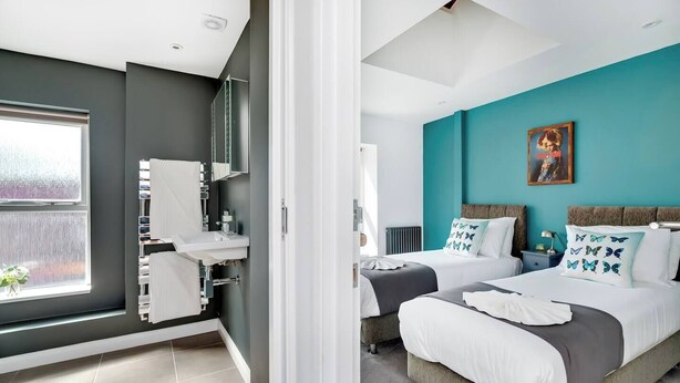 Habitación de hotel moderna con un ventanal grande, un baño con lavamanos y toallero, y dos camas con almohadas con motivos de mariposas.