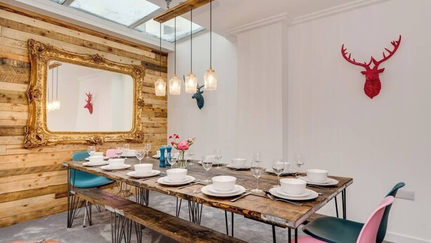 Un comedor con una mesa de madera dispuesta para seis comensales, un espejo grande y una decoración con cabeza de ciervo en la pared.