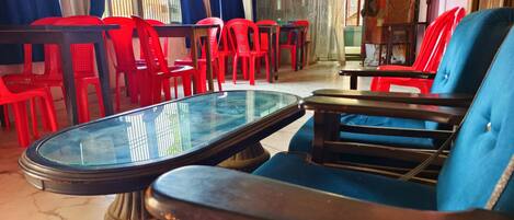 A room with a glass-top table and blue chairs, red plastic chairs, and a wooden table.