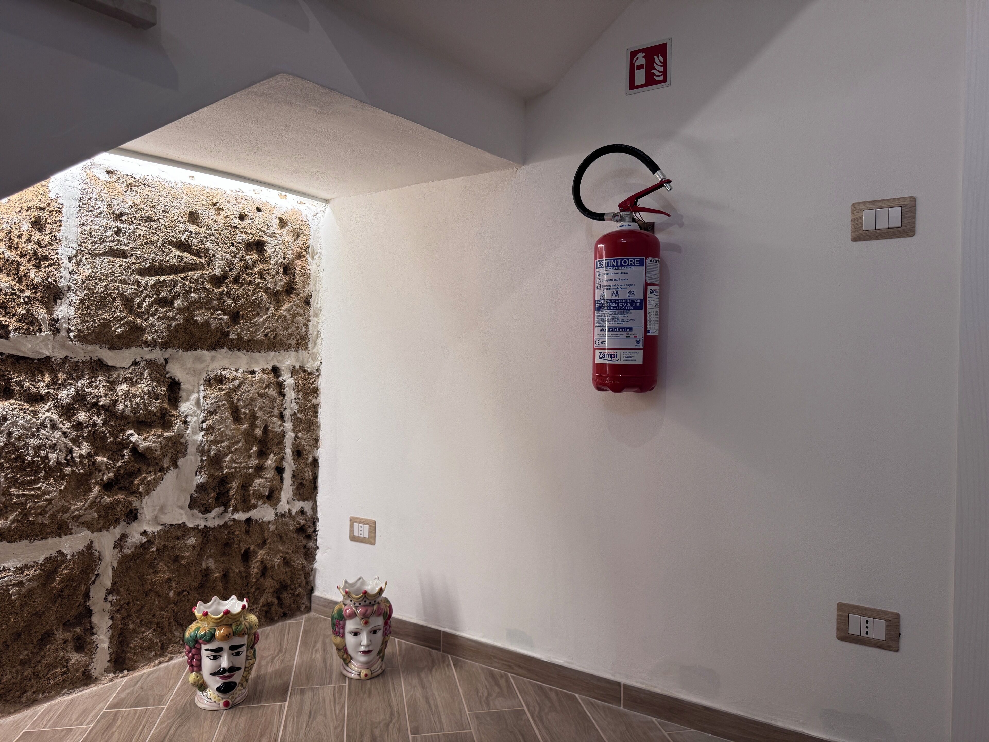Una habitación con pared de piedra, un extintor de incendios sujeto a la pared y una cabeza de cerámica decorativa en el suelo.