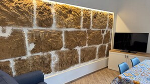 A modern living room with a stone wall, a flat-screen TV, a sofa, and a dining area with a patterned tablecloth. - Dimora del Tufo Antico - House in the heart of Avola (Avola)