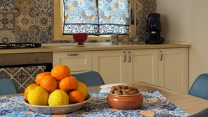 A kitchen with a gas stove, a coffee maker, a window with patterned curtains, and a table with a bowl of oranges and lemons. - Dimora del Tufo Antico - House in the heart of Avola (Avola)
