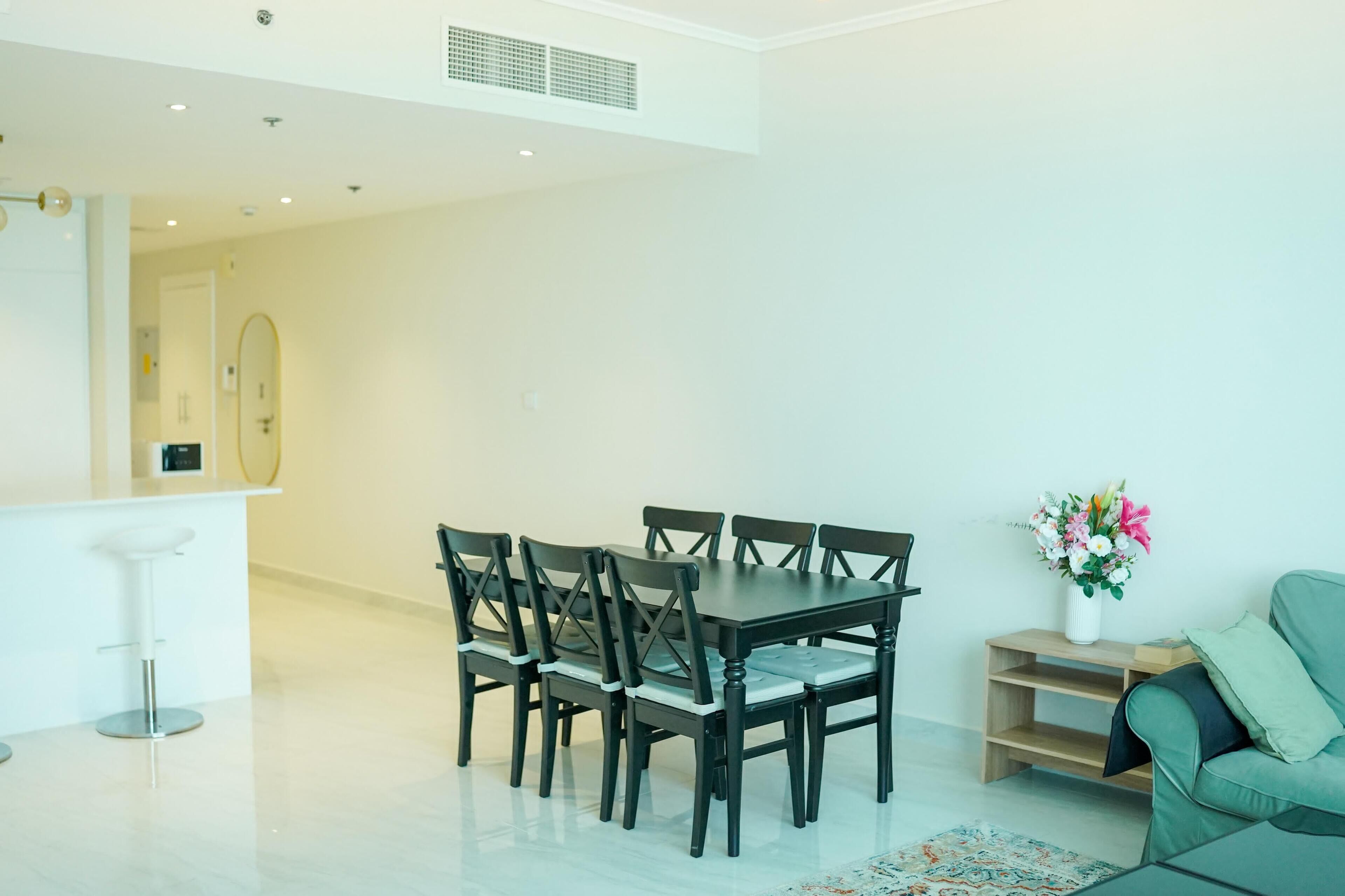 A modern living room with a dining table and chairs, a green sofa, a small wooden side table with a vase of flowers, and a white bar area with a stool.