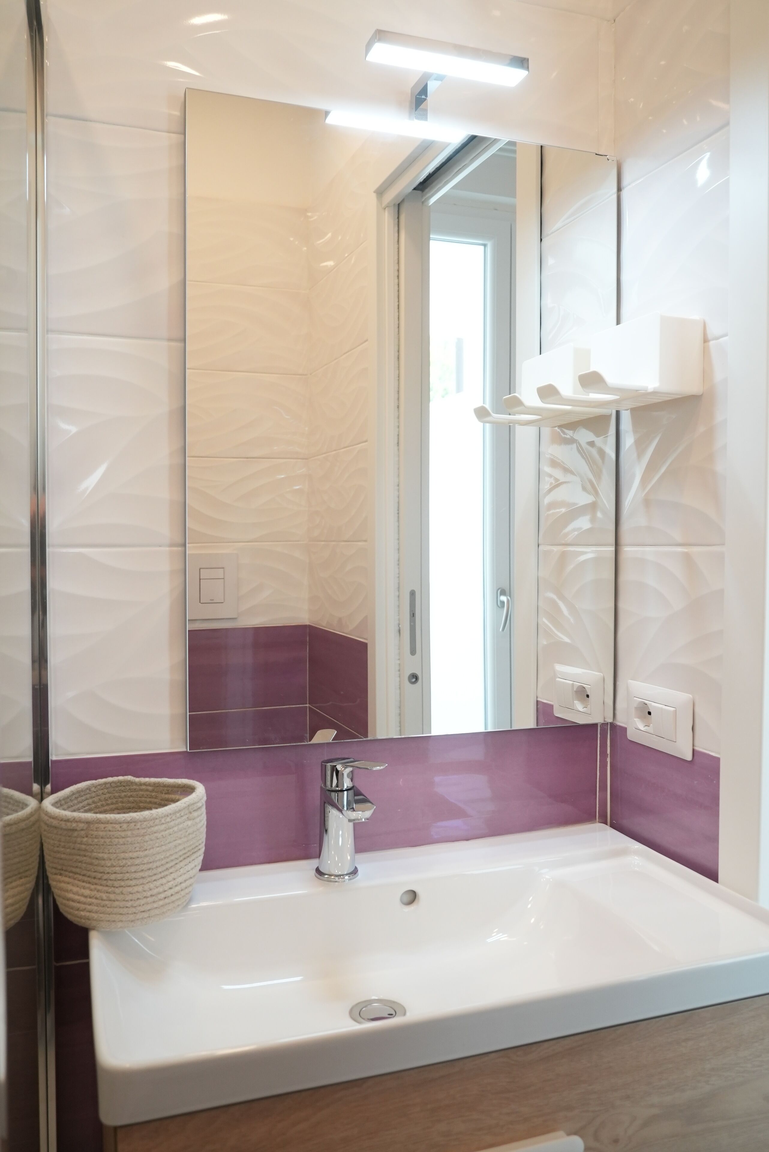 A modern bathroom with a large mirror, white sink, and purple-tiled walls.