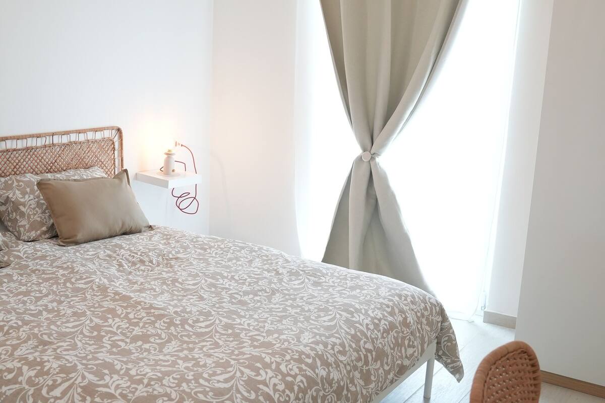 A bedroom with a large bed, a bedside table with a lamp, a wicker chair, and a window with curtains.