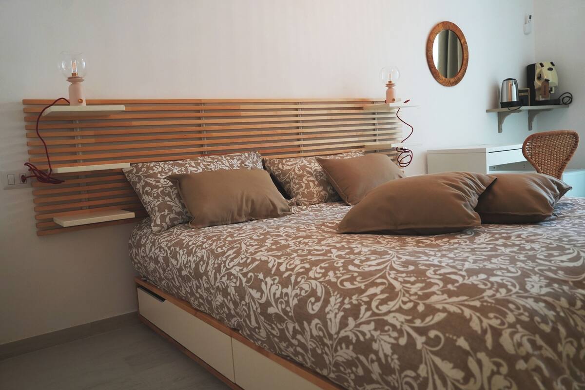 A bedroom with a bed, wooden headboard, bedside table, lamp, mirror, and a chair.