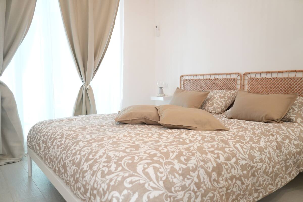 A neatly made bed with a patterned bedspread, a bedside table with a lamp, and a window with curtains.