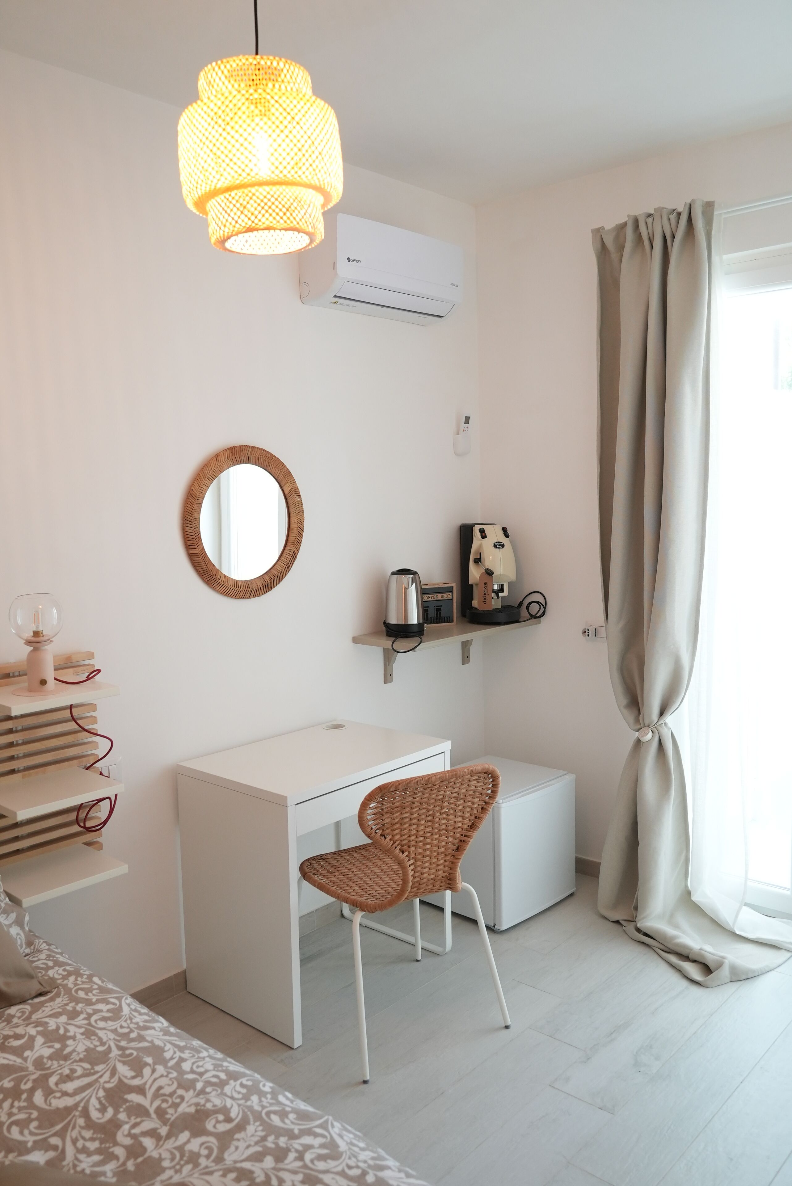 Un dormitorio moderno con un escritorio blanco, una silla de mimbre, un espejo redondo y una lámpara colgante.