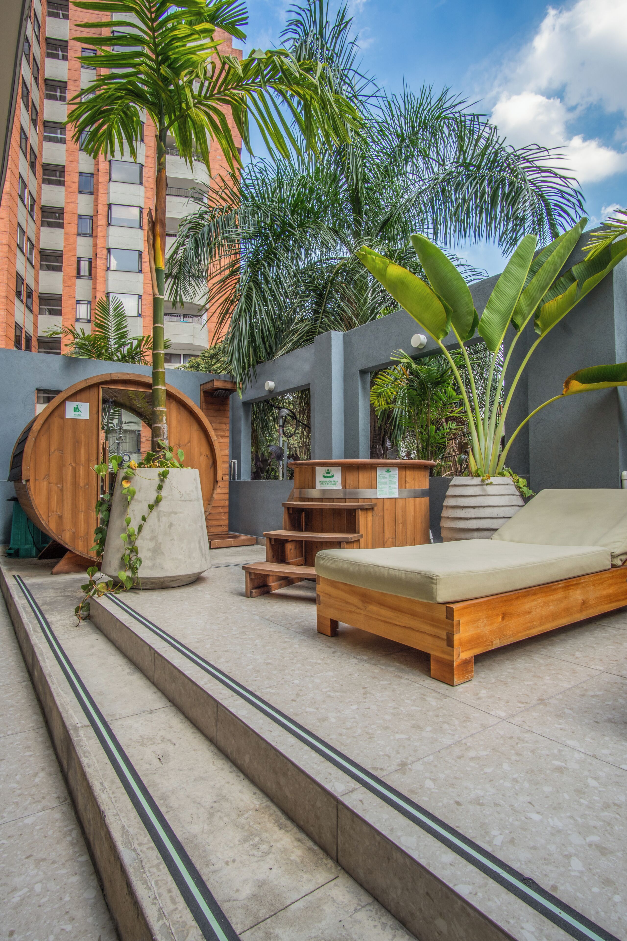 A wooden bench, a lounge chair, a wooden barrel, and a potted plant in an outdoor area with steps leading up to a building.